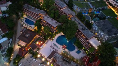 Deniz kıyısındaki akşam kenti. Yukarıdan bak. Gecenin ışıkları parlıyor, arabalar ve scooterlar geçiyor. Havuzda ışıklar parlıyor. Evler yeşil tepelerde yer alıyor. Phuket