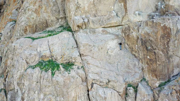 A man climbs a rock. Active recreation and sports. Mountaineering with equipment. Extreme outdoor activities. High rocks of Tamgaly TAS, Kazakhstan. Top view from a drone. Green grass.
