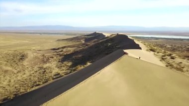 Kazakistan 'ın büyük kum tepeleri. Şarkı söyleyen bir kumulun üstündeki bir dronun en üst görüntüsü. Bir grup insan bir dağın tepesinde dinleniyor. Bir sürü sarı kum ve insan izi var. Bulutlu mavi gökyüzü