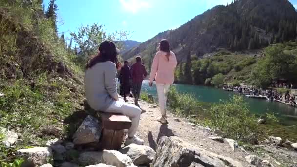男が湖沿いのリュックを持って歩いている 人々は ベンチに座ってリラックスし 山の湖コルセーでボートに乗る 野外活動 歩道を歩いてください モミの木が成長します カザフスタン — ストック動画