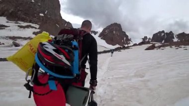 Büyük bir sırt çantasıyla zirveye tırmanıyor. Bir dağcı karda dağlık bir alanda yürüyor. İyi çiğnenmiş bir yol, büyük kayalar ve kasvetli bir gökyüzü. Gökyüzündeki bulutlar. Ayakaltında kirli kar.