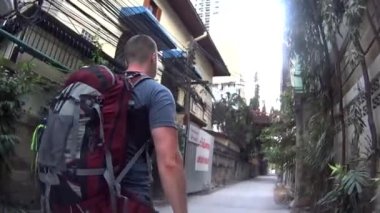 Büyük sırt çantalı bir çift şehirde dolaşıyorlar. Sokakta turist bir kızla yürüyen adam. Turistlerin arkasında kocaman sırt çantaları var. Uzun evleri olan küçük bir sokak. Yeşil bitkiler her yerde yetişir.