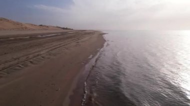 Dawn on the sandy beach of the sea. Sand dune. The suns rays peek out from behind the clouds. Car tracks in the sand. A light wave, almost calm. The water merges with the sky. Autumn morning