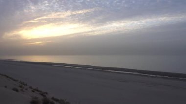 Dawn on the sandy beach of the sea. Sand dune. The suns rays peek out from behind the clouds. Car tracks in the sand. A light wave, almost calm. The water merges with the sky. Autumn morning