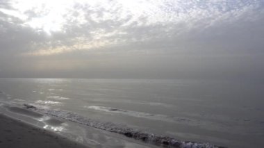 Dawn on the sandy beach of the sea. Sand dune. The suns rays peek out from behind the clouds. Car tracks in the sand. A light wave, almost calm. The water merges with the sky. Autumn morning