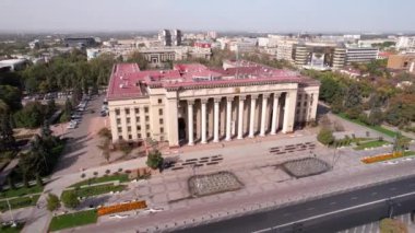 Aerial view of the old square of Almaty. A beautiful building resembling a palace. Nearby fountains, a park and a road. The flag of Kazakhstan on the roof of the Kazakh-British Technical University.