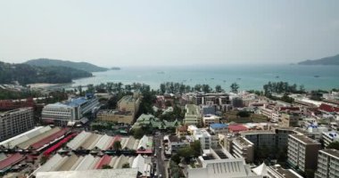 Patong, Phuket adasının insansız hava aracı görüntüsü. Sahilde farklı yükseklikteki evler duruyor. Adanın yeşil tepeleri. Evlerin havuzlarında insanlar yüzer. Yolda scooter 'lar ve arabalar.