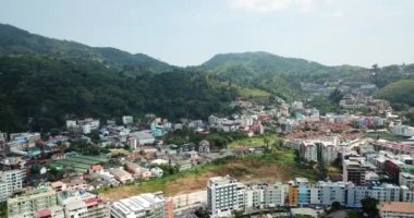 Patong, Phuket adasının insansız hava aracı görüntüsü. Sahilde farklı yükseklikteki evler duruyor. Adanın yeşil tepeleri. Evlerin havuzlarında insanlar yüzer. Yolda scooter 'lar ve arabalar.
