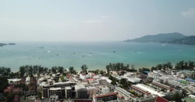 Patong, Phuket adasının insansız hava aracı görüntüsü. Sahilde farklı yükseklikteki evler duruyor. Adanın yeşil tepeleri. Evlerin havuzlarında insanlar yüzer. Yolda scooter 'lar ve arabalar.