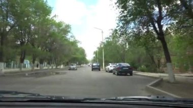 View of the hood of the car and the broken road. There are a lot of pits, wells and cracks on the asphalt. The car is driving through city. Green trees grow, there are small houses. Sky with clouds