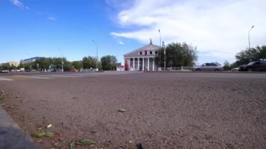 Arabalar ve insanlarla kesişme zamanı. Mavi gökyüzünde beyaz bulutlar hızlı uçuyor. Saray şeklinde yeşil ağaçlar ve bir bina var. Trafik ışıkları yanıp sönüyor. Balkhash şehri