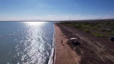 Mavi renkli ve kumlu dalgalı bir sahil. Tatil yapan insanların, çadırların ve arabaların görüntüsü. Balkhash Gölü 'nün kumları. Mavi gökyüzü. Çalılıklar ve birkaç ağaç yetişiyor. Deniz kenarında aile tatili