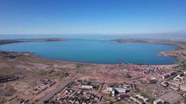 Drone view of Bertys Bay, Lake Balkhash. The blue-green water of the lake reflects the sky and clouds. There is a town and a factory on the shore. The coastal line is clearly visible, low houses