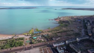 Drone view of Bertys Bay, Lake Balkhash. The blue-green water of the lake reflects the sky and clouds. There is a town and a factory on the shore. The coastal line is clearly visible, low houses