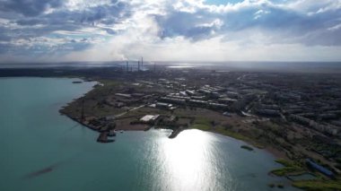 Drone view of Bertys Bay, Lake Balkhash. The blue-green water of the lake reflects the sky and clouds. There is a town and a factory on the shore. The coastal line is clearly visible, low houses