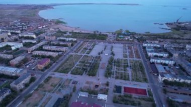 Aerial view from a drone of the small town of Balkhash on the shore of the lake. Low houses are painted in different colors, wide alleys, green trees grow. Cars are driving along the road. The clouds