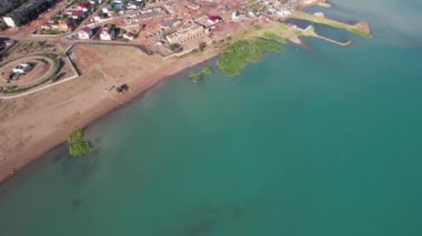 Drone view of Bertys Bay, Lake Balkhash. The blue-green water of the lake reflects the sky and clouds. There is a town and a factory on the shore. The coastal line is clearly visible, low houses
