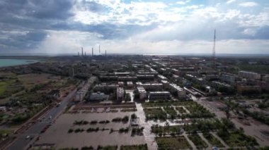 Raindrops fall on the city and the smoking factory. Blue-white clouds float over the city. There is a tall TV tower. Cars drive along the roads, people walk along the alley. Drone view of Balkhash