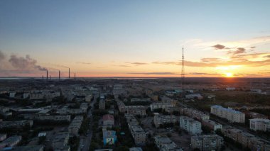 Orange sunset over a small town. Top view from a drone. Blue-purple clouds against background of houses, a smoking factory and a TV tower. Cars drive. There is a bright light at the metal drain plant