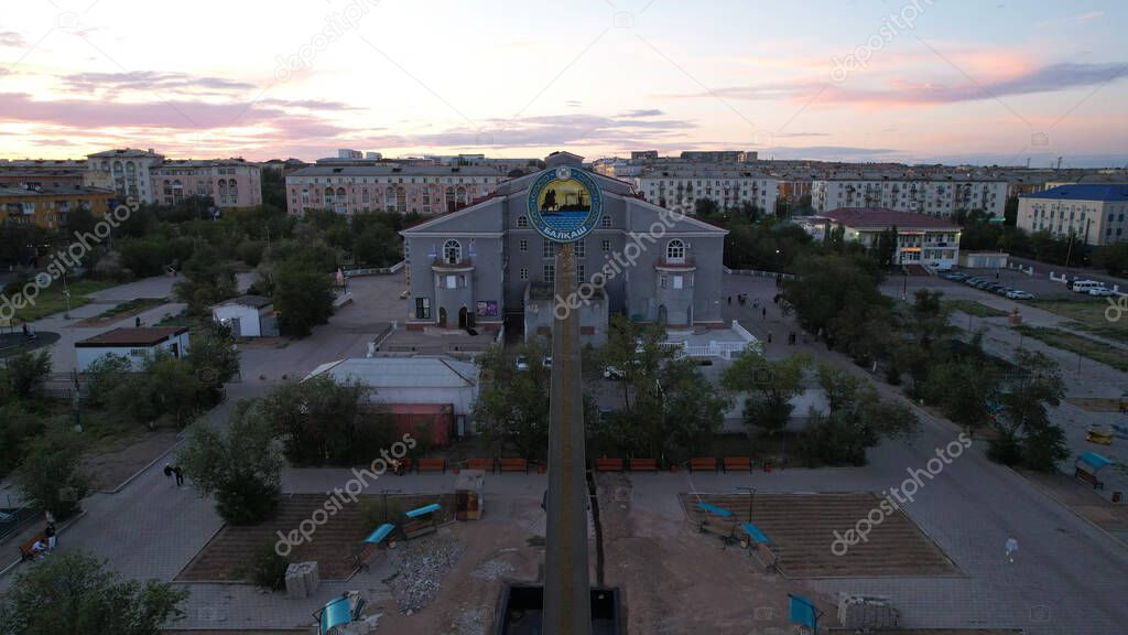 Monument to city of Balkhash and metallurgists at sunset. The fountain is being reconstructed. Pink sky with clouds. Low houses and a palace. People walk along green alley. Flower beds with flowers