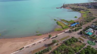 Drone view of Bertys Bay, Lake Balkhash. The blue-green water of the lake reflects the sky and clouds. There is a town and a factory on the shore. The coastal line is clearly visible, low houses