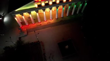 Bright color illumination of a beautiful palace. The night sky with the moon. People are walking along the alley, sitting on benches. Reflection in the water of the fountain. Tall columns of building