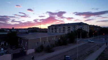 Pink sunset over a small town. Top view from a drone. Flocks of swifts fly. The sun goes below horizon. Blue-purple clouds on background of houses, a factory and a TV tower. Cars drive, people rest