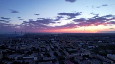Pink sunset over a small town. Top view from a drone. Flocks of swifts fly. The sun goes below horizon. Blue-purple clouds on background of houses, a factory and a TV tower. Cars drive, people rest