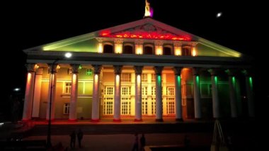 Bright color illumination of a beautiful palace. The night sky with the moon. People are walking along the alley, sitting on benches. Reflection in the water of the fountain. Tall columns of building