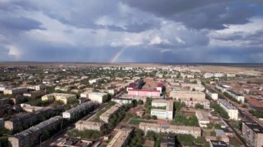 Bright rainbow and rain over the city of Balkhash. The small town is all wet. Large clouds of white-blue color float by. Cars drive and people walk along the streets. Green trees are growing