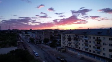 Pink sunset over a small town. Top view from a drone. Flocks of swifts fly. The sun goes below horizon. Blue-purple clouds on background of houses, a factory and a TV tower. Cars drive, people rest
