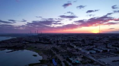 Top view from a drone. The sun is sinking below the horizon. Blue-purple clouds on the background of houses, factories and TV towers. Cars are driving, people are resting. Bertys Bay, Lake Balkhash