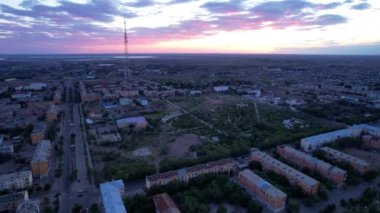 Pink sunset over a small town. Top view from a drone. Flocks of swifts fly. The sun goes below horizon. Blue-purple clouds on background of houses, a factory and a TV tower. Cars drive, people rest
