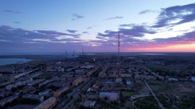 Pink sunset over a small town. Top view from a drone. Blue-purple clouds against background of houses, a smoking factory and a TV tower. Cars drive. There is a bright light at the metal drain plant