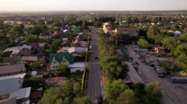 A black car is driving on a road in a small town. There are a lot of green trees and bushes. Low houses of different colors. A straight road. Top view from the drone on the highway.