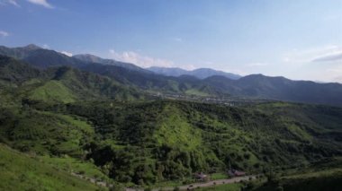 The mountains are covered with green trees and grasses. Top view from the drone on the road with cars, there are houses in places. A small town is visible in the distance. A green forest is growing