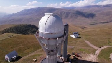 The Assy-Turgen Observatory with a large telescope. A tent camp is located nearby. There are colored tents, cars and people are resting. Big clouds over green hills. Two domes for studying the stars