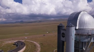 The Assy-Turgen Observatory with a large telescope. A tent camp is located nearby. There are colored tents, cars and people are resting. Big clouds over green hills. Two domes for studying the stars