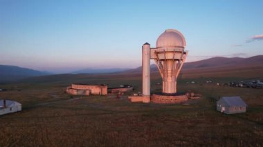 Two large telescope domes at sunset. Drone view of Assy-Turgen Observatory. Beautiful red sunset. Green hills and clouds. Tourists watch the sun. There is a large tent camp and cars nearby. Kazakhstan