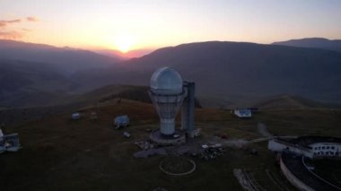 Two large telescope domes at sunset. Drone view of Assy-Turgen Observatory. Beautiful red sunset. Green hills and clouds. Tourists watch the sun. There is a large tent camp and cars nearby. Kazakhstan