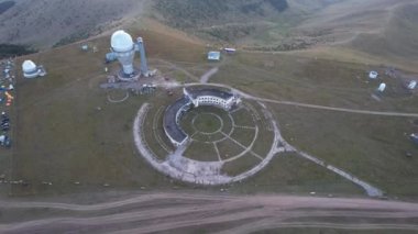 Two large telescope domes at sunset. Drone view of Assy-Turgen Observatory. Beautiful red sunset. Green hills and clouds. Tourists watch the sun. There is a large tent camp and cars nearby. Kazakhstan