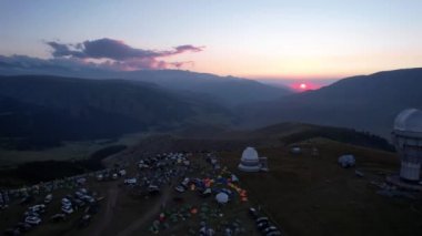 Two large telescope domes at sunset. Drone view of Assy-Turgen Observatory. Beautiful red sunset. Green hills and clouds. Tourists watch the sun. There is a large tent camp and cars nearby. Kazakhstan