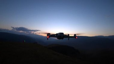 A DJI Air2S drone with red-green lights hovers above the ground. Sunset is being filmed. The sun goes behind the mountains. Clouds and snowy peaks are visible in the distance. Tourists admire sunset