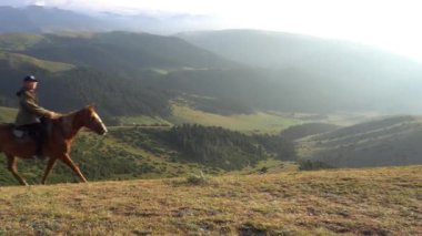 A guy in a cap rides a horse on a yellow-green hill. A young horse follows nature. Mountains, clouds, green forests and large bushes are visible in the distance. Steep hills are covered with forests