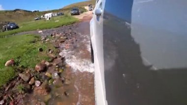 The jeep drives along a dirt road across river. Splashes from the wheels fly in different directions. There are small stones. Green fields, hills and forest are visible. People are relaxing in nature