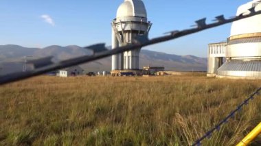 A large observatory is protected by a barbed fence. There is a long sharp wire around perimeter. Buildings with domes and a telescope are visible in distance. Yellow-green grass. Mountainous terrain