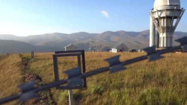 A large observatory is protected by a barbed fence. There is a long sharp wire around perimeter. Buildings with domes and a telescope are visible in distance. Yellow-green grass. Mountainous terrain