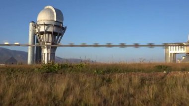 A large observatory is protected by a barbed fence. There is a long sharp wire around perimeter. Buildings with domes and a telescope are visible in distance. Yellow-green grass. Mountainous terrain
