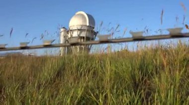 A large observatory is protected by a barbed fence. There is a long sharp wire around perimeter. Buildings with domes and a telescope are visible in distance. Yellow-green grass. Mountainous terrain