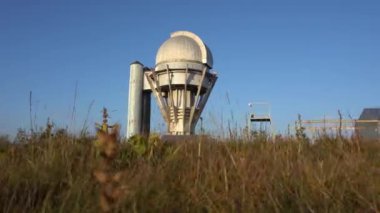 Large buildings of the observatory in the form of a dome. The telescopes are closed. Beautiful tall yellow-green grass develops in the wind. Blue sky and green hills in the distance. The Assy Plateau